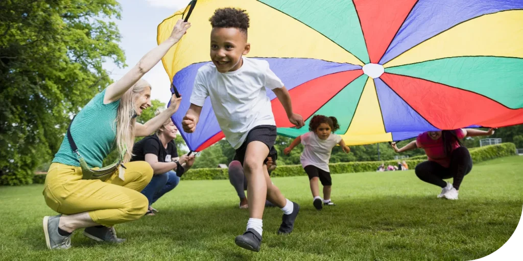 children at summer school laughing and playing with a large, colorful parachute, lifting it into the air together as part of a group activity.